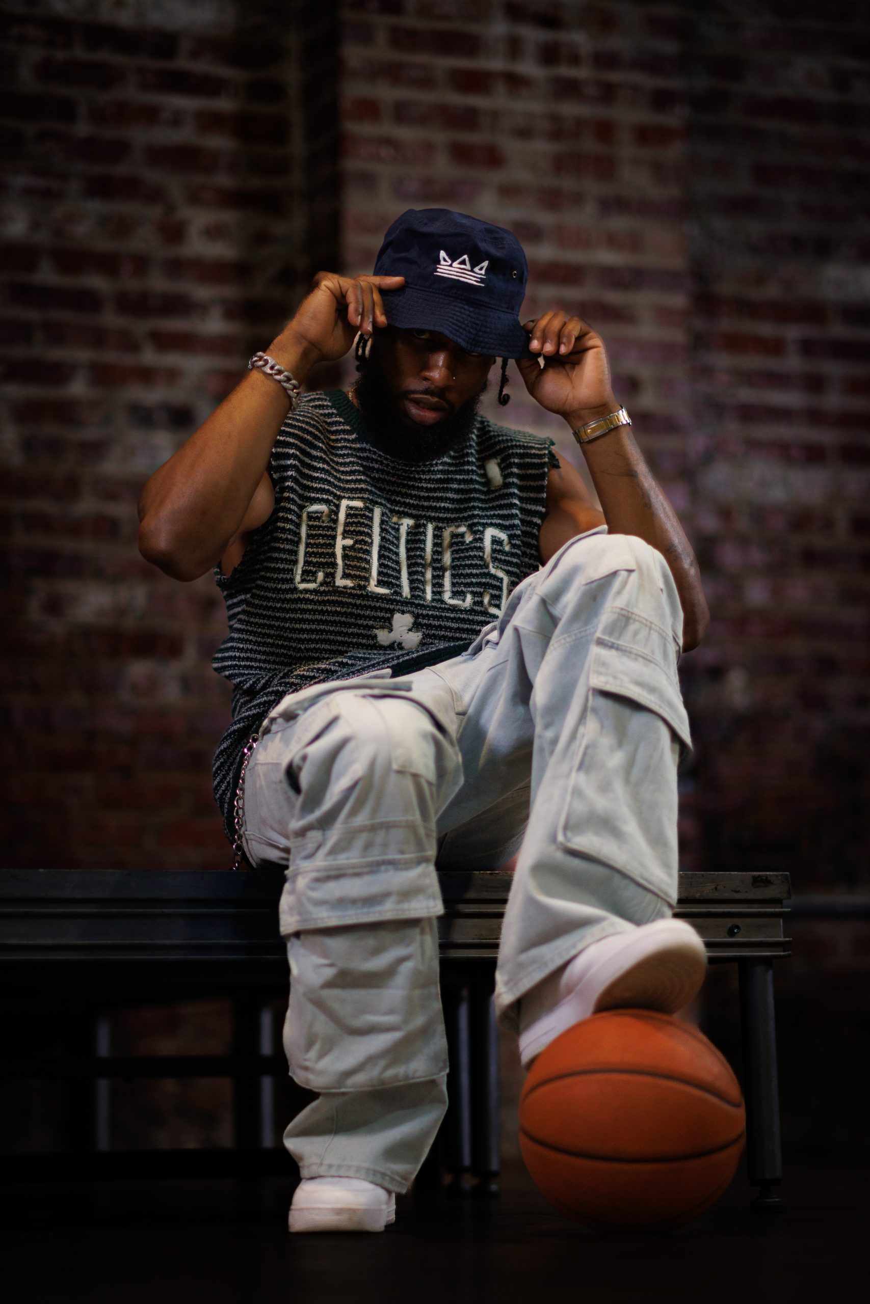 Man in a Celtics jersey adjusting his bucket hat, seated with a basketball