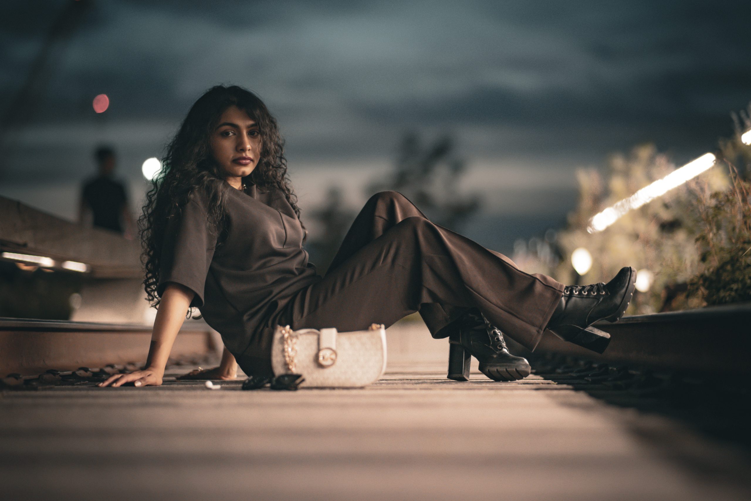 Portrait photographer's editorial shot: woman with long curly hair reclining on railway tracks at dusk, string lights in the background