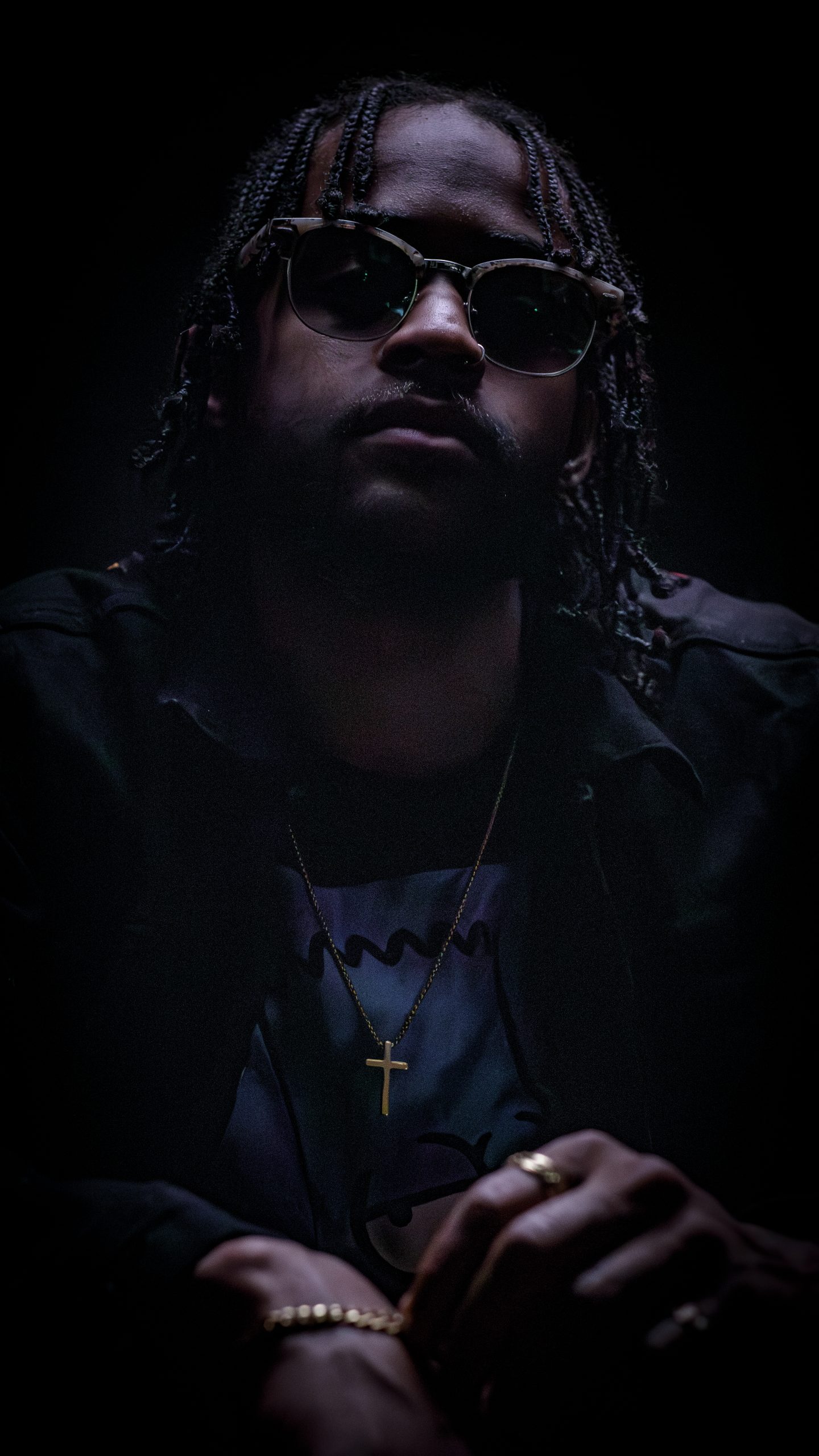 Man with braided hair and sunglasses in a dark, moody portrait, gold cross necklace